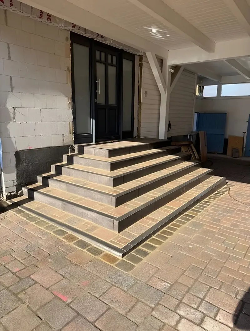 Über Eck laufende Treppe aus Blockstufen vor einem Hauseingang, davor eine Pflasterfläche