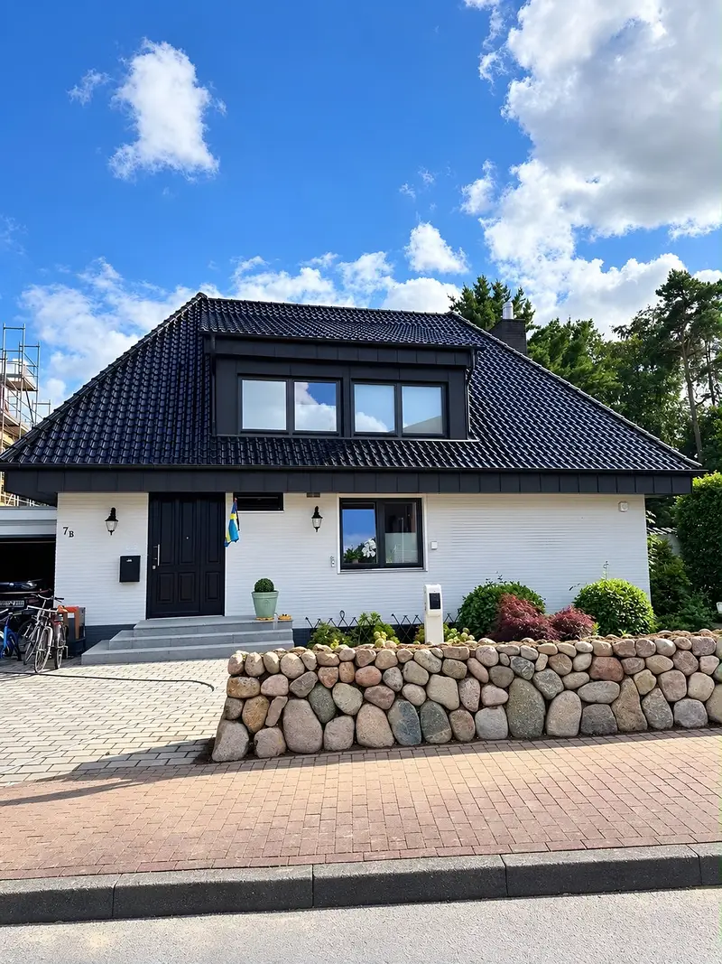 Friesenwall aus runden Feldsteinen an der Grundstücksgrenze eines weißen Einfamilienhauses, davor ein Gehweg aus rotem Klinker