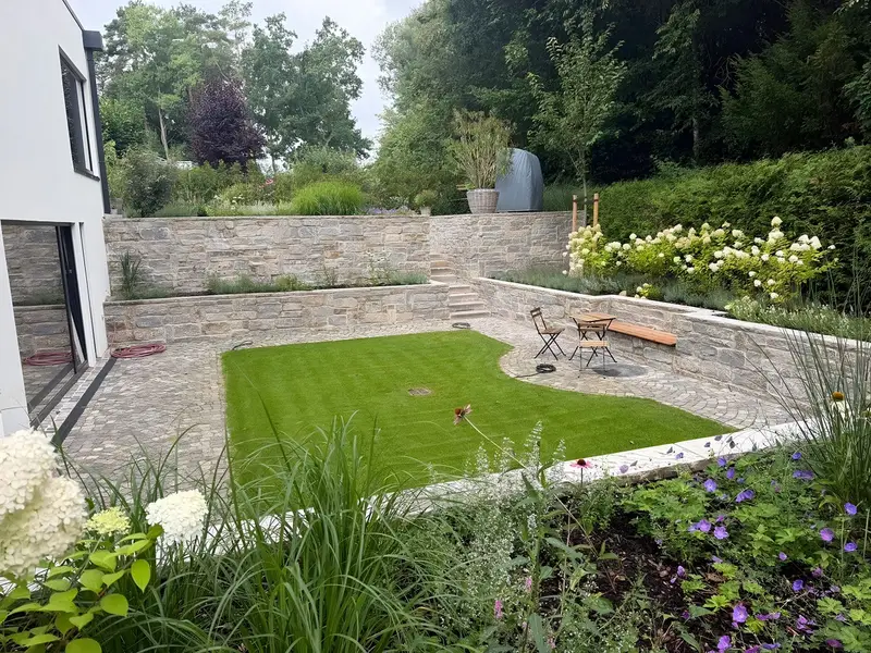 Garten mit geschwungener Rasenfläche, gepflasterter Sitzecke, Natursteinmauer und bepflanzten Beeten