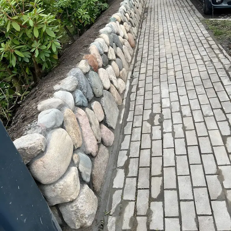 Gehweg aus hellem Klinker, daneben ein Friesenwall aus großen runden Feldsteinen mit bepflanzter Krone