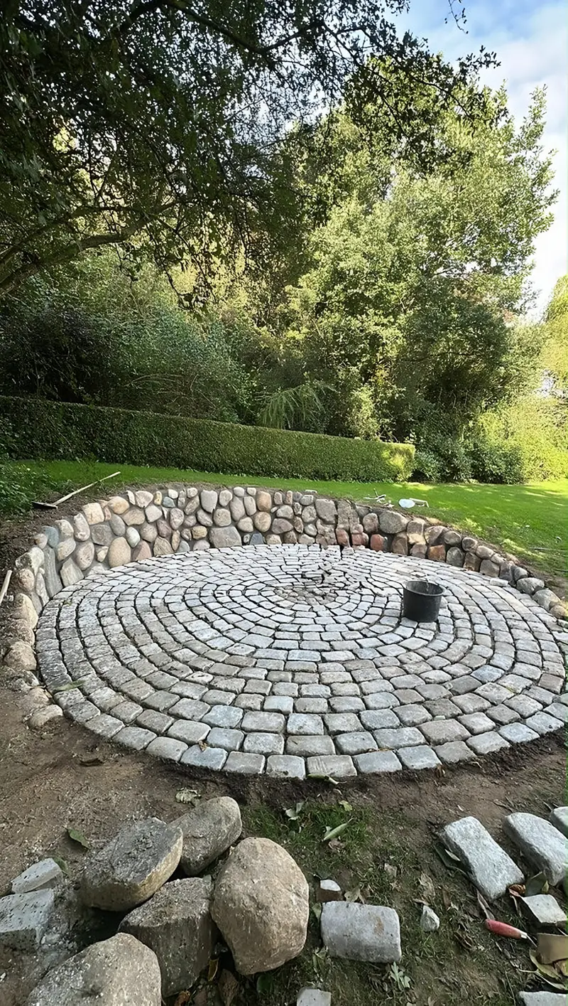 Kreisrunder Platz aus Natursteinpflaster in einem Garten, eingefasst von einer niedrigen Feldsteinmauer